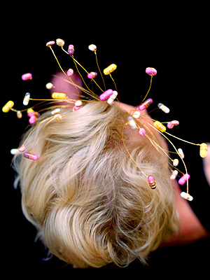 A blonde woman's head from above, wearing a tiara wreathed in yellow, pink and white pills.